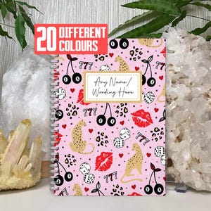 May include: A pink spiral-bound notebook featuring a pattern of cherries, dice, leopard print, and lips. The notebook has a white label with "Any Name/Wording Here" and a red banner that says "20 Different Colours".