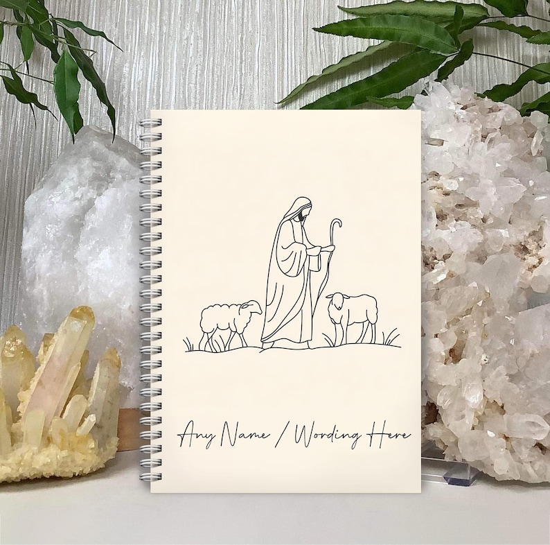 May include: A cream-colored spiral notebook with a black line drawing of a shepherd with two sheep. The text "Any Name / Wording Here" is at the bottom of the notebook.