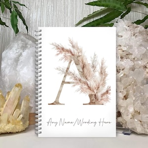 May include: White spiral-bound notebook with a large letter "A" decorated with a watercolour pampas grass design in beige and brown tones. The text "Any Name/Wording Here" is written in a script font at the bottom of the notebook.