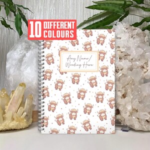May include: A spiral-bound notebook with a white cover featuring a repeating pattern of cartoon Highland cows in shades of brown and beige. The notebook has a rectangular label with the text "Any Name/Wording Here". A red rectangle reads "10 DIFFERENT COLOURS".