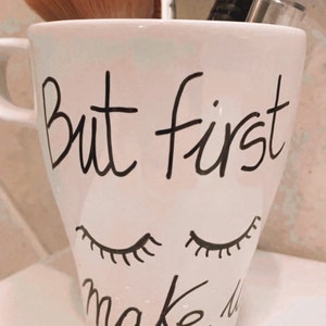 Può includere: Una tazza in ceramica bianca con il testo "But first make up" scritto in nero. La tazza ha due ciglia nere disegnate sul davanti. Un pennello per il trucco e uno strumento argentato sono nella tazza.