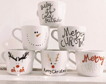 May include: A collection of white ceramic mugs with Christmas-themed designs. Some mugs have text like "Baby it's cold outside" and "Merry Christmas." Others feature festive illustrations of a snowman and reindeer. The mugs are arranged on a white shelf.