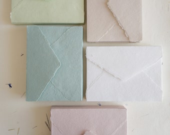 May include: Five stacks of handmade paper envelopes in different colors: light green, light blue, white, light pink, and beige. The envelopes have a textured, handmade look and are all the same size.