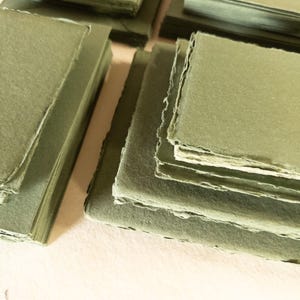 May include: Stacks of handmade, olive green paper products. The image shows several stacks of square paper sheets and folded envelopes, all in a muted green hue. The edges of the paper have a rough, uneven texture.