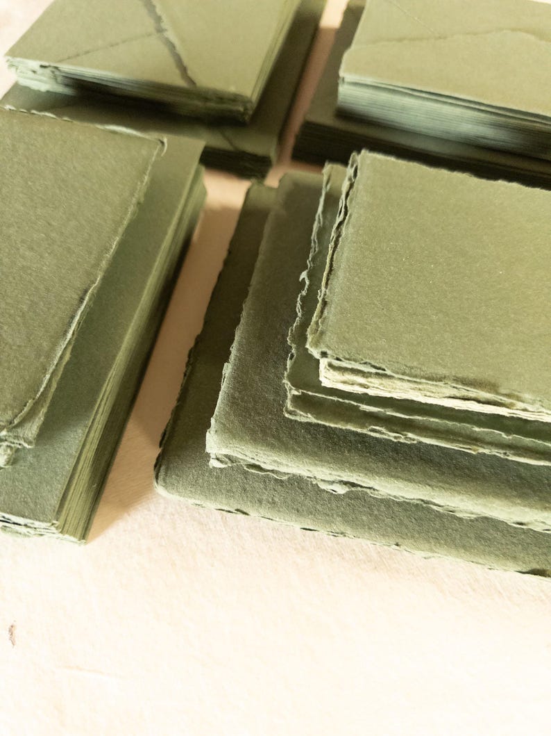 May include: Stacks of handmade, olive green paper products. The image shows several stacks of square paper sheets and folded envelopes, all in a muted green hue. The edges of the paper have a rough, uneven texture.