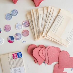 Könnte beinhalten: Eine Anordnung aus handgefertigten Papierherzen in verschiedenen Größen und Rosatönen. Wachssiegel mit Monogrammen, Buchseiten und ein kleiner Umschlag mit Briefmarken und Adressinformationen sind ebenfalls vorhanden.