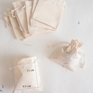 Puede incluir: Un conjunto de 10 pequeñas bolsas de algodón con cordón de color blanco roto. Las bolsas están apiladas unas sobre otras. Una sola bolsa se muestra por separado con medidas de 9,5 cm por 11 cm.