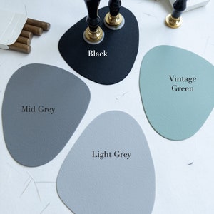 May include: Four different coloured leather coasters, in black, mid grey, light grey, and vintage green. The coasters are shaped like rounded rectangles. The black coaster has two black and gold wax seal stamps on top of it.