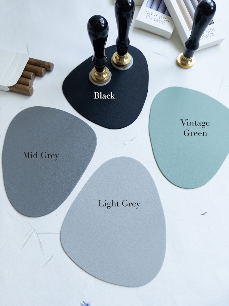 May include: Four different coloured leather coasters, in black, mid grey, light grey, and vintage green. The coasters are shaped like rounded rectangles. The black coaster has two black and gold wax seal stamps on top of it.