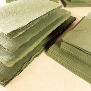 May include: Stacks of handmade, olive-green paper cards with rough, deckled edges. The cards are arranged in several piles, some neatly stacked and others slightly fanned out. The paper has a textured appearance, suggesting a natural fiber composition.