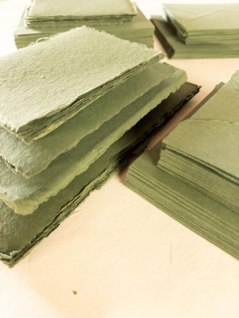 May include: Stacks of handmade, olive-green paper cards with rough, deckled edges. The cards are arranged in several piles, some neatly stacked and others slightly fanned out. The paper has a textured appearance, suggesting a natural fiber composition.
