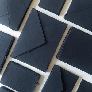 May include: A collection of black paper envelopes with a rough, textured finish. The envelopes are stacked and arranged in a pattern on a white surface.