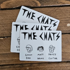 May include: Three white rectangular stickers with the band name "THE CHATS" in bold, black, jagged lettering. Below the band name are three cartoon illustrations with the names "Eamon Bass", "Matt Drums", and "Pricey Guitar". The stickers are on a wooden surface.