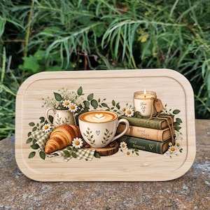 Könnte beinhalten: Ein rechteckiges Serviertablett aus Bambus mit einem aufgedruckten Motiv aus einer Kaffeetasse mit Latte Art, einem Croissant auf einer karierten Serviette, einem kleinen Gänseblümchenkrug und drei Büchern, auf denen eine Kerze steht.