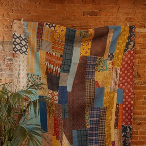 May include: A colorful patchwork quilt with a variety of fabrics and patterns in shades of blue, yellow, red, and brown. The quilt is hanging on a brick wall.