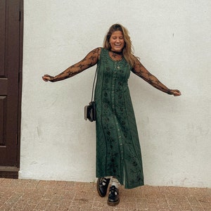 Pode incluir: Uma mulher vestindo um vestido maxi verde com mangas de renda preta e sapatos pretos. Ela está em pé em frente a uma parede branca com uma porta marrom.