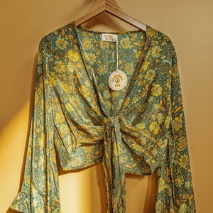 May include: A green and yellow floral print blouse with a tie front and long bell sleeves. The blouse has a V-neck and is made of a lightweight fabric. The tag on the blouse reads "Gardenella Shop".