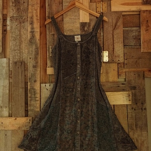 Könnte beinhalten: Ein dunkelbraunes, ärmelloses Kleid mit Knopfleiste und Blumenmuster. Das Kleid hängt an einem Holzbügel vor einem rustikalen Holzhintergrund.