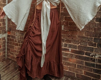 May include: A woman wearing a white peasant top with a brown skirt and long brown braids. The top is tied at the front and has long, flowing sleeves. The skirt is gathered at the waist and has a ruffled hem. The woman is standing in front of a brick wall.