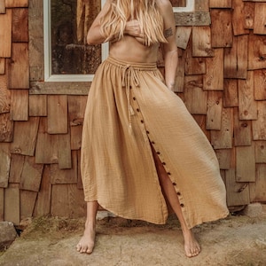 May include: A woman wearing a long, flowing, light brown skirt with a button-up front and a drawstring waist. She is standing in front of a wooden building with a window behind her.