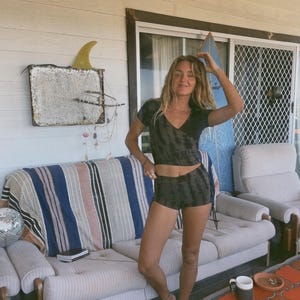 May include: A woman wearing a black and green tie-dye crop top and matching shorts. The woman is standing on a wooden deck in front of a white couch with a striped blanket. A disco ball and a surfboard are visible.