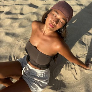 May include: A person wearing a brown tube top, light gray shorts, and a maroon headscarf, sitting on a sandy beach. The person is wearing silver hoop earrings. The image is taken in natural sunlight.