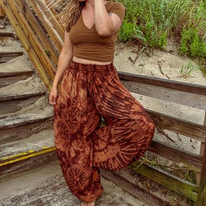 May include: Brown tie-dye harem pants with a matching brown V-neck crop top. The pants feature a unique circular and striped pattern. The outfit is worn on a wooden boardwalk near the beach.