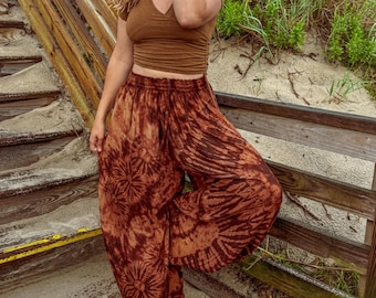 Può includere: Pantaloni harem marroni con stampa tie-dye e un top corto marrone con scollo a V abbinato. I pantaloni presentano un motivo circolare e a righe unico. L'outfit è indossato su una passerella di legno vicino alla spiaggia.