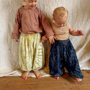 May include: Two children wearing colorful tie-dye pants. The child on the left is wearing a long-sleeved, mauve shirt and green and yellow tie-dye pants. The child on the right is wearing a striped tank top and dark blue tie-dye pants.