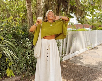 May include: A woman wearing a green cropped top and a white maxi skirt with button detailing. She is holding a cup of coffee and smiling. The background features a tree with Spanish moss hanging from its branches.