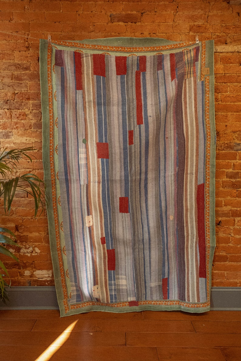 Op de afbeelding: Een vintage gestreept tapijt met een groene rand en rode, blauwe en bruine strepen. Het tapijt hangt aan een bakstenen muur.