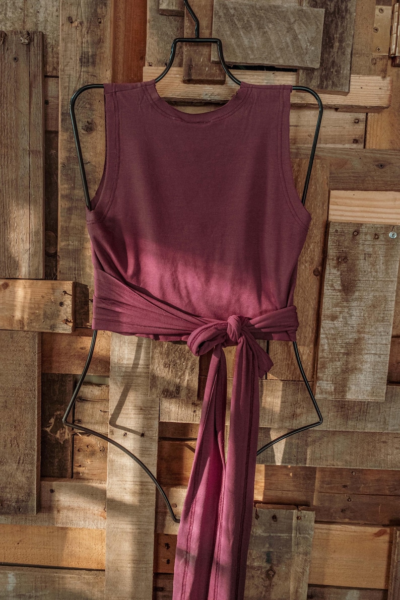 May include: A maroon sleeveless top with a tie at the waist. The top is hanging on a black hanger against a wooden background.