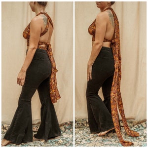 May include: A woman is wearing a black halter top with a colorful patterned fabric tied around her waist and a long scarf. She is also wearing black flared jeans.