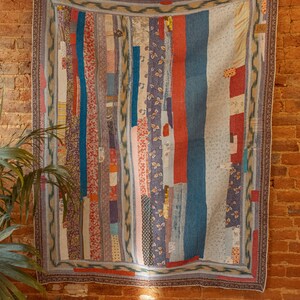 Op de afbeelding: Een patchworkdeken met verschillende kleuren en patronen, waaronder blauw, rood, roze en wit. De deken hangt aan een bakstenen muur.