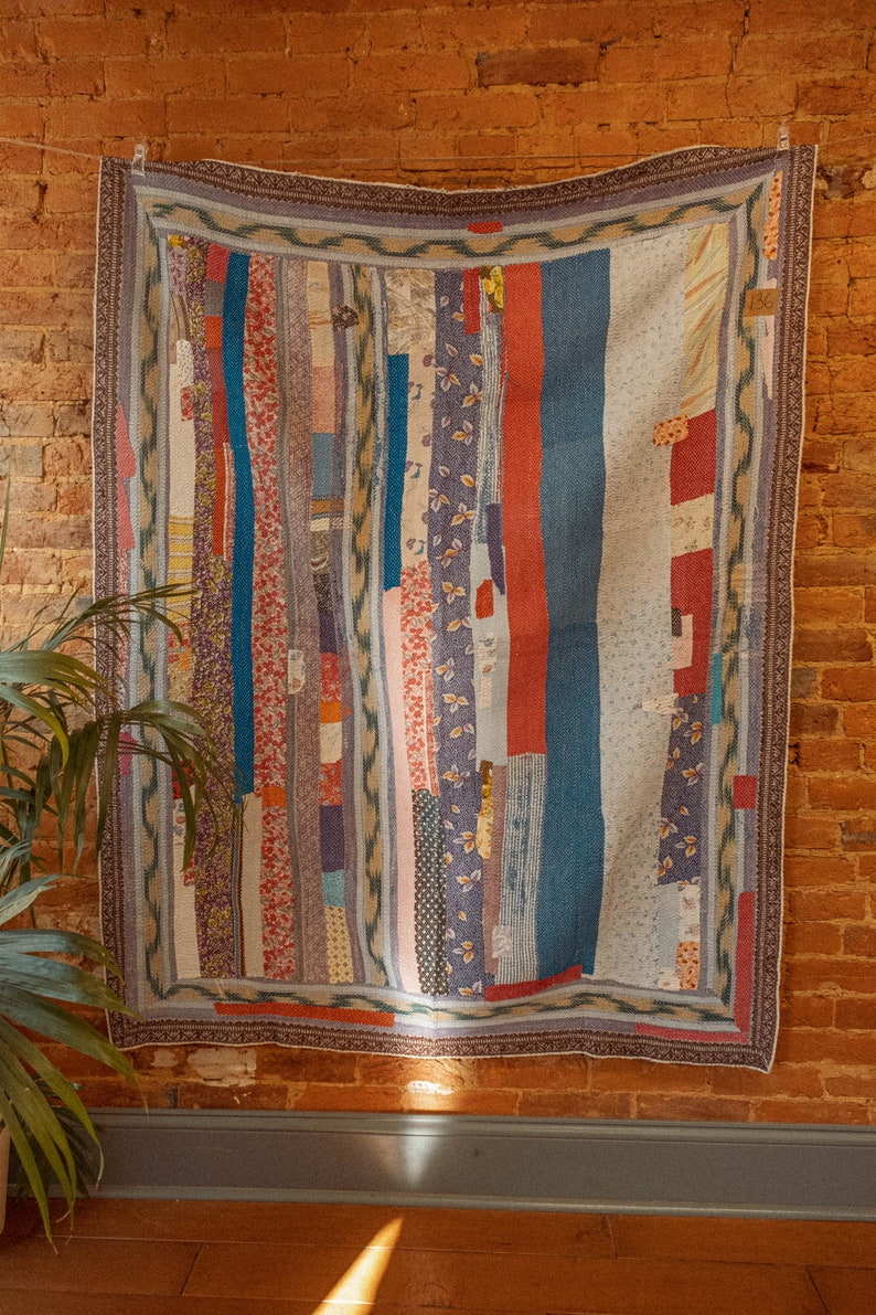 Op de afbeelding: Een patchworkdeken met verschillende kleuren en patronen, waaronder blauw, rood, roze en wit. De deken hangt aan een bakstenen muur.
