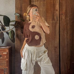 May include: A brown and beige yin-yang design crop top and off-white, loose-fitting cargo trousers. The top has a round neckline and short sleeves. The trousers have pockets and a drawstring waist. The outfit is casual and comfortable.