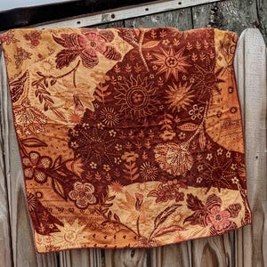 Puede incluir: Un textil cuadrado con un estampado floral en tonos marr&oacute;n, naranja y beige. La tela est&aacute; drapeada sobre una superficie de madera, mostrando los intrincados detalles del estampado bot&aacute;nico. El textil presenta varios motivos de flores y rayos de sol.
