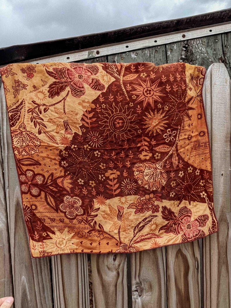 Puede incluir: Un textil cuadrado con un estampado floral en tonos marr&oacute;n, naranja y beige. La tela est&aacute; drapeada sobre una superficie de madera, mostrando los intrincados detalles del estampado bot&aacute;nico. El textil presenta varios motivos de flores y rayos de sol.