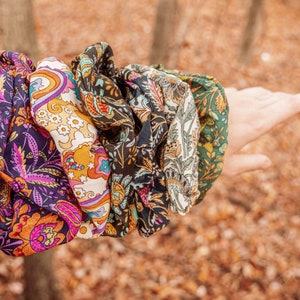 May include: A set of five colorful patterned hair scrunchies. The scrunchies are made of fabric with a paisley design and are arranged in a stack on a person's arm.