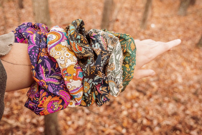 May include: A set of five colorful patterned hair scrunchies. The scrunchies are made of fabric with a paisley design and are arranged in a stack on a person's arm.