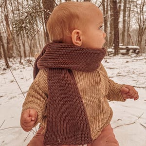 May include: A toddler wearing a brown knitted scarf, a beige knitted sweater, and pink pants sits in the snow. The child is looking to the right of the frame.