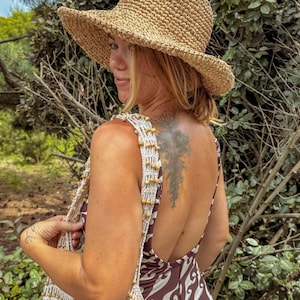 Pu&ograve; includere: Un cappello da sole in paglia intrecciata con tesa larga. Il cappello &egrave; color beige e ha una corona leggermente arrotondata. La persona indossa un vestito fantasia marrone e bianco e una borsa intrecciata bianca e beige con perline di legno.