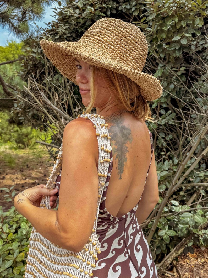 Pu&ograve; includere: Un cappello da sole in paglia intrecciata con tesa larga. Il cappello &egrave; color beige e ha una corona leggermente arrotondata. La persona indossa un vestito fantasia marrone e bianco e una borsa intrecciata bianca e beige con perline di legno.