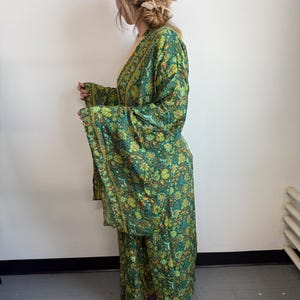 Puede incluir: Un kimono verde con estampado floral con mangas anchas y una falda a juego. La tela presenta un patrón de flores y hojas amarillas y naranjas sobre un fondo verde. La prenda la lleva una persona.