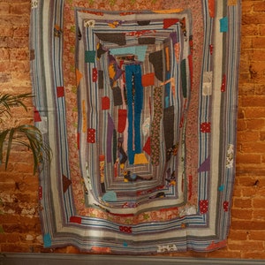 Op de afbeelding: Een kleurrijk patchwork-deken met een geometrisch ontwerp. Het deken is gemaakt van verschillende stoffen in tinten blauw, rood, groen en geel. Het deken hangt aan een bakstenen muur.