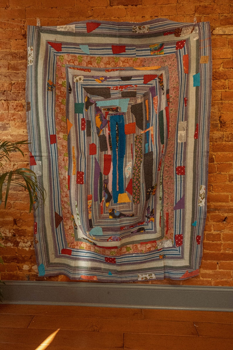 Op de afbeelding: Een kleurrijk patchwork-deken met een geometrisch ontwerp. Het deken is gemaakt van verschillende stoffen in tinten blauw, rood, groen en geel. Het deken hangt aan een bakstenen muur.
