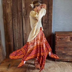 May include: A cream-colored long-sleeve sweater paired with a flowing, handkerchief-hem skirt featuring a vibrant, multi-colored floral and geometric print in shades of orange, red, and pink, worn with brown leather boots.