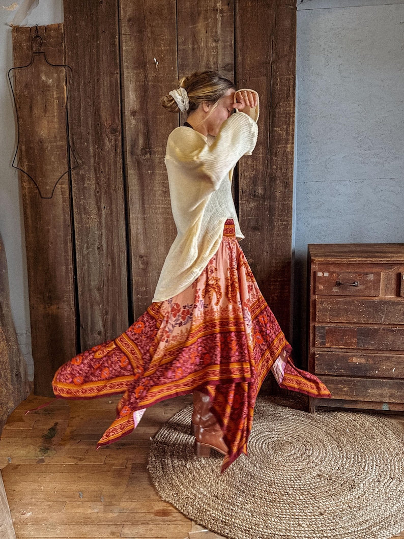 May include: A cream-colored long-sleeve sweater paired with a flowing, handkerchief-hem skirt featuring a vibrant, multi-colored floral and geometric print in shades of orange, red, and pink, worn with brown leather boots.