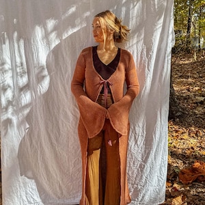 May include: A long, open-front rust-colored knit cardigan with bell sleeves. The cardigan is paired with matching pants and a dark brown top. The outfit is tied together with a ribbon. The background is a white sheet and a forest.