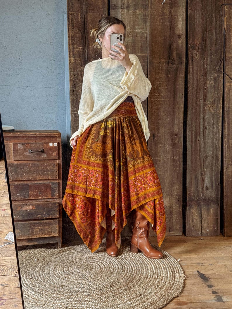 May include: A cream-colored, long-sleeved, lightweight knit sweater paired with a brown, orange, and yellow patterned midi skirt featuring a handkerchief hem, an elasticized waistband, and intricate floral and animal motifs, worn with brown leather boots.
